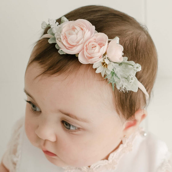 A close-up photo of a baby wearing a Joli Flower Headband, featuring pink roses and green leaves, while dressed in a white lace outfit. The baby gazes downward, showcasing dark eyelashes.