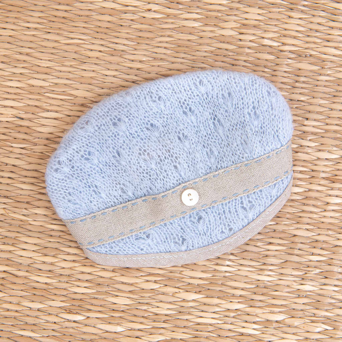 A light blue oval-shaped Austin Hat with a beige strap secured with a small white button, placed on a woven straw-like surface.