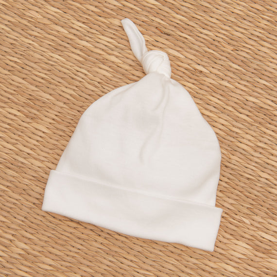 A white Mason Knot Cap lies flat on a textured tan wicker background. The hat has a soft texture and a playful top knot, perfect for a christening.