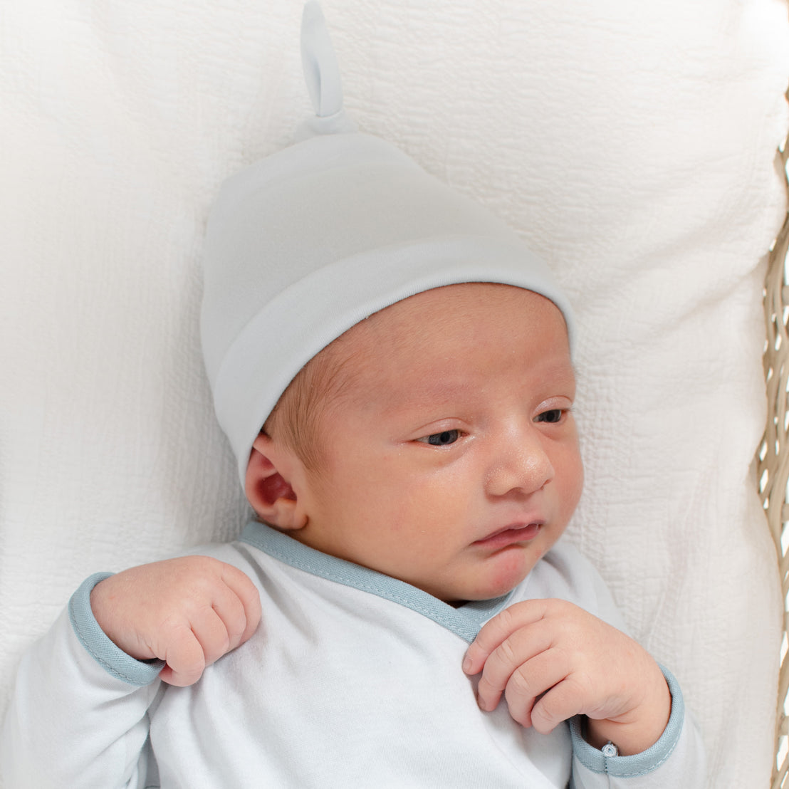 A newborn baby with a light complexion, wearing an Aiden Newborn Knot Cap and a blue outfit, lying comfortably in a white basket, looking slightly to the side as they are coming home from the hospital.