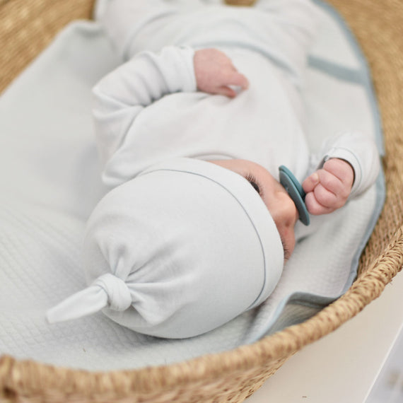 A newborn baby swaddled in a light blue outfit with an Aiden Newborn Knot Cap, sleeping peacefully in a boutique woven basket. The basket is lined with soft white fabric, perfect for coming home from the hospital.