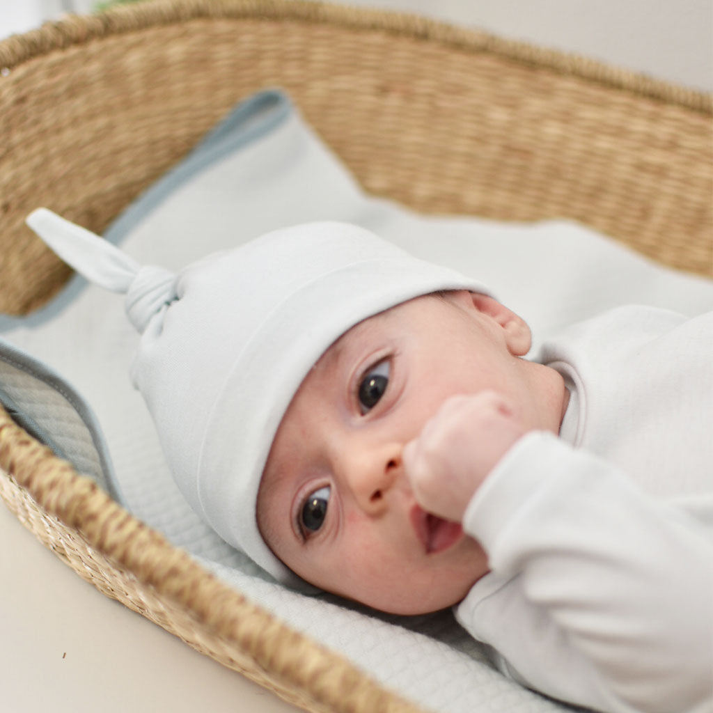 A newborn lies in an upscale wicker basket, wearing a white onesie and an Aiden Newborn Knot Cap with a pointed tip. The baby gazes upward with one hand near the mouth.
