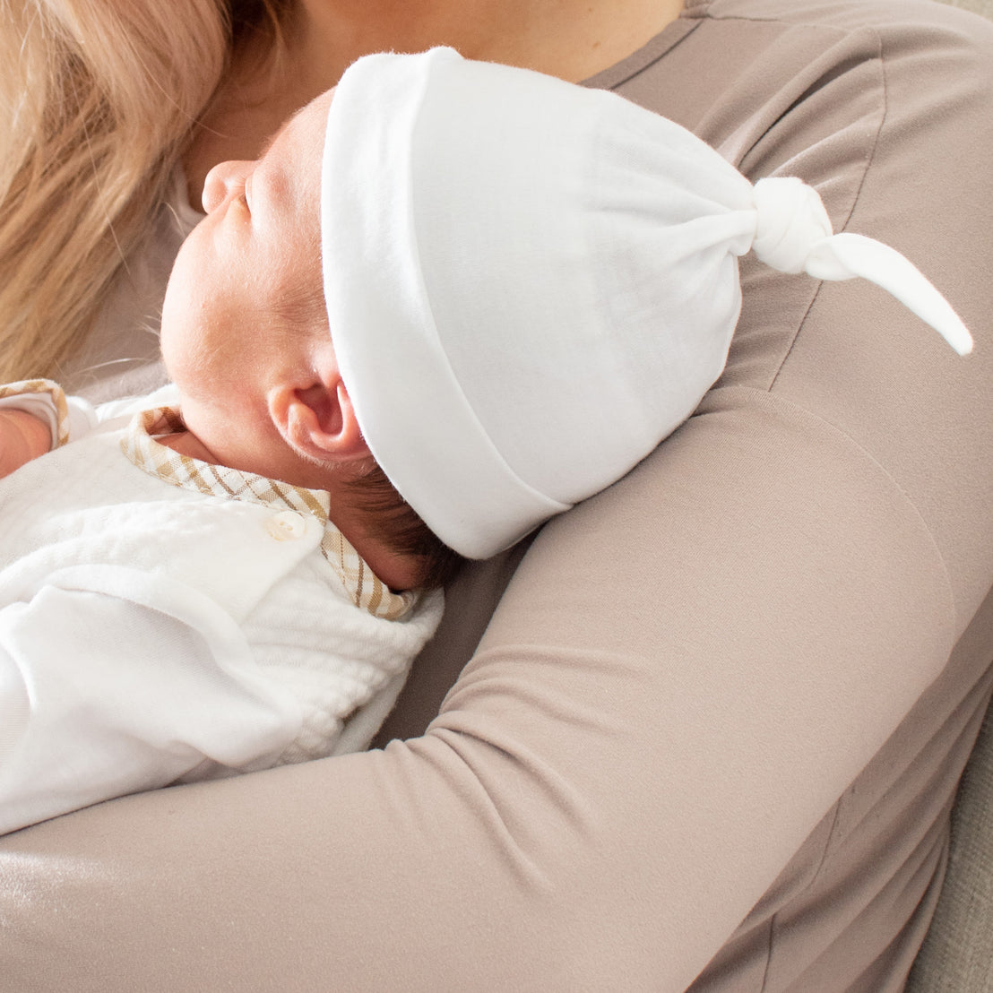 A close-up image of a newborn sleeping peacefully on a woman's shoulder. The baby wears a white Dylan Knot Cap, including a hat, and is wrapped in a white blanket.