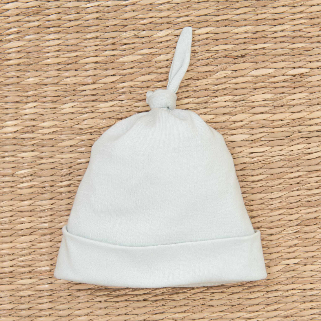 A plain white Aiden Newborn Knot Cap, laid flat on a textured beige woven background.