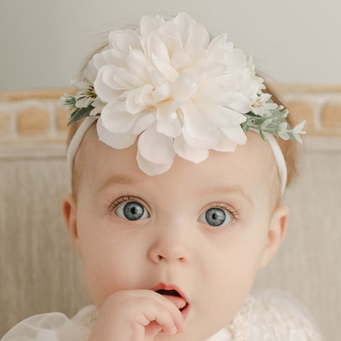 A baby with striking blue eyes looks in surprise or curiosity during her christening, adorned with a large white Kristina Flower Headband.