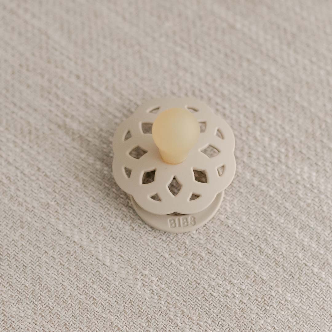 A vintage-inspired Vanilla & Ivory pacifier with an intricate pattern and the brand name "bibs" on a textured grey fabric background.