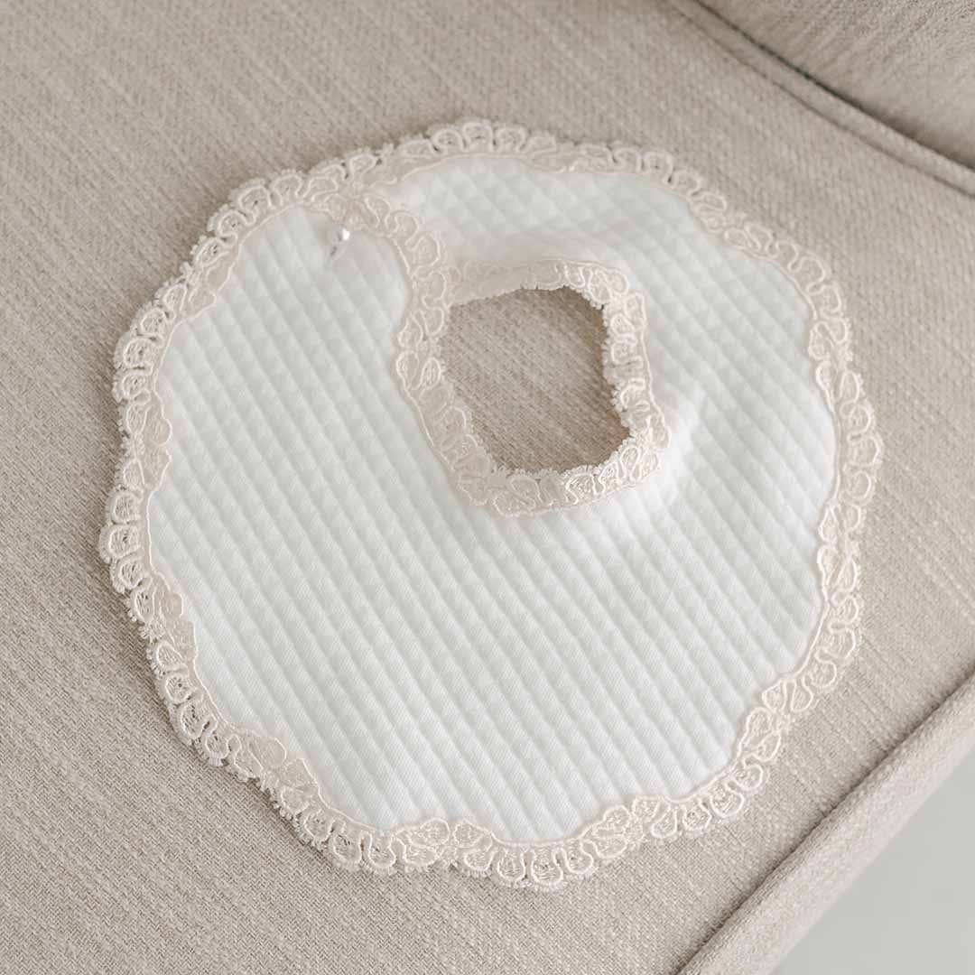 A vintage-inspired white quilted Kristina Bib with lace trim, perfect for a baptism or christening, displayed on a beige fabric surface.