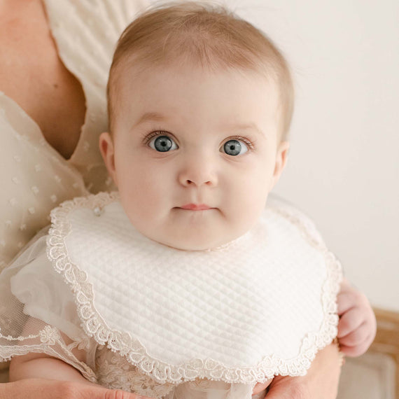 Baby with wide blue eyes and a white Kristina Bib looks curiously at the camera during a christening.