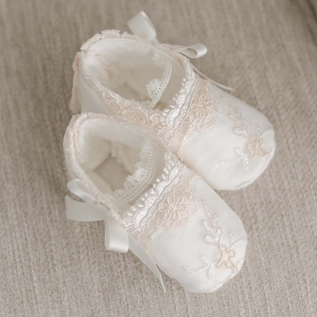 A pair of delicate white Kristina Booties with intricate lace and embroidery details, tied with satin ribbons for a baptism, displayed on a soft beige fabric background.