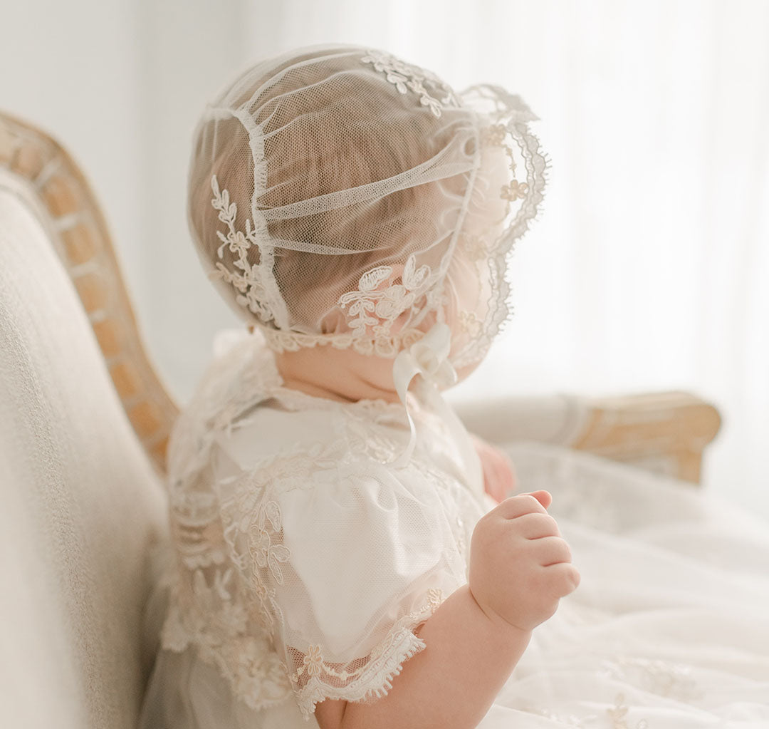 A baby wearing a Kristina Christening Gown & Bonnet with lace details and a matching bonnet sits on an elegant chair, the bonnet's veil partially covering the head, in a softly lit room.