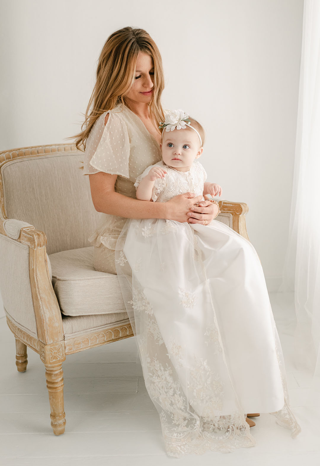 A woman sits on an elegant armchair holding a baby dressed in a white lace gown and headband for a christening, both looking towards the camera in a bright room