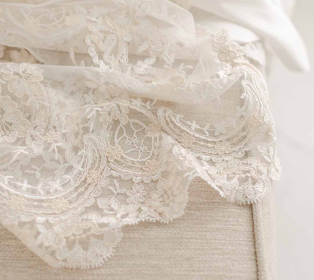 Close-up of delicate white Kristina Christening Gown & Bonnet lace fabric with intricate floral patterns, draped softly over a neutral-toned surface, creating a gentle and elegant texture perfect for a christening.