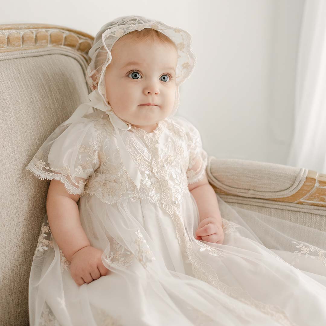 A baby with blue eyes sitting on a beige sofa, wearing the Kristina Christening Gown & Bonnet, looking up with a curious expression.