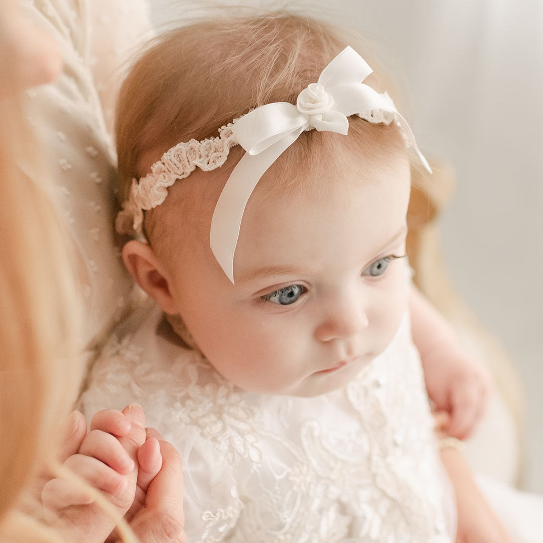 A close-up of a baby with blue eyes and a Kristina Headband, held by an adult wearing a vintage-inspired lace dress. The focus is on the baby's contemplative expression.