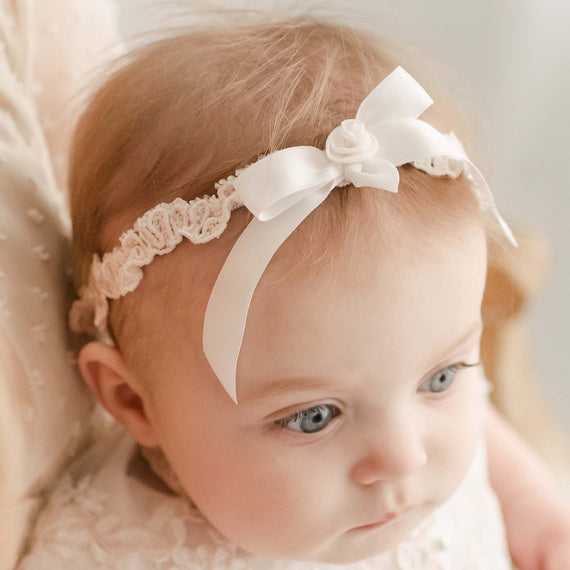 Close-up of a baby with blue eyes and light hair, wearing a white lace Kristina headband with a bow, looking thoughtfully to the side.