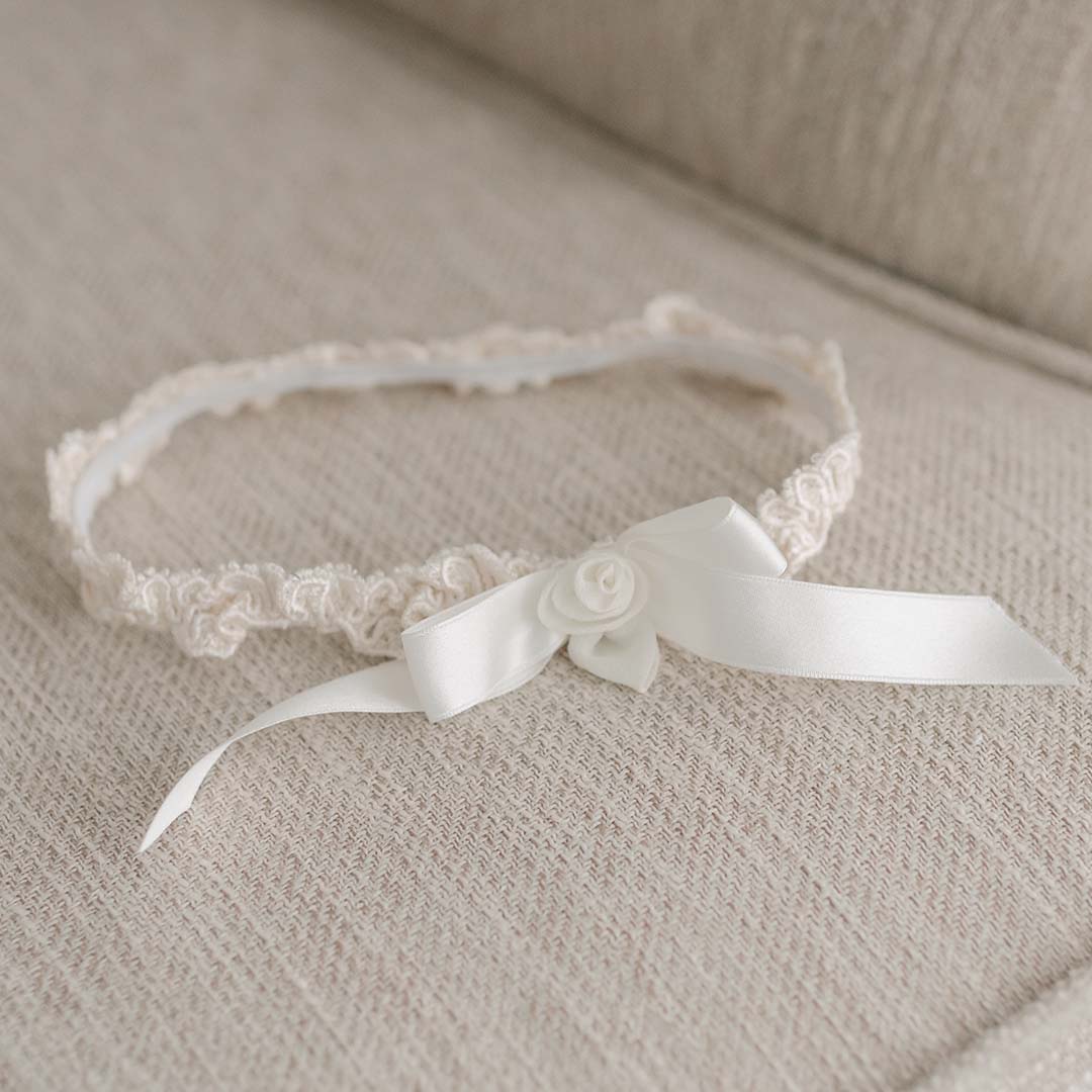 A delicate white lace Kristina Headband with a small satin bow and a rose detail, displayed on a soft beige fabric background.