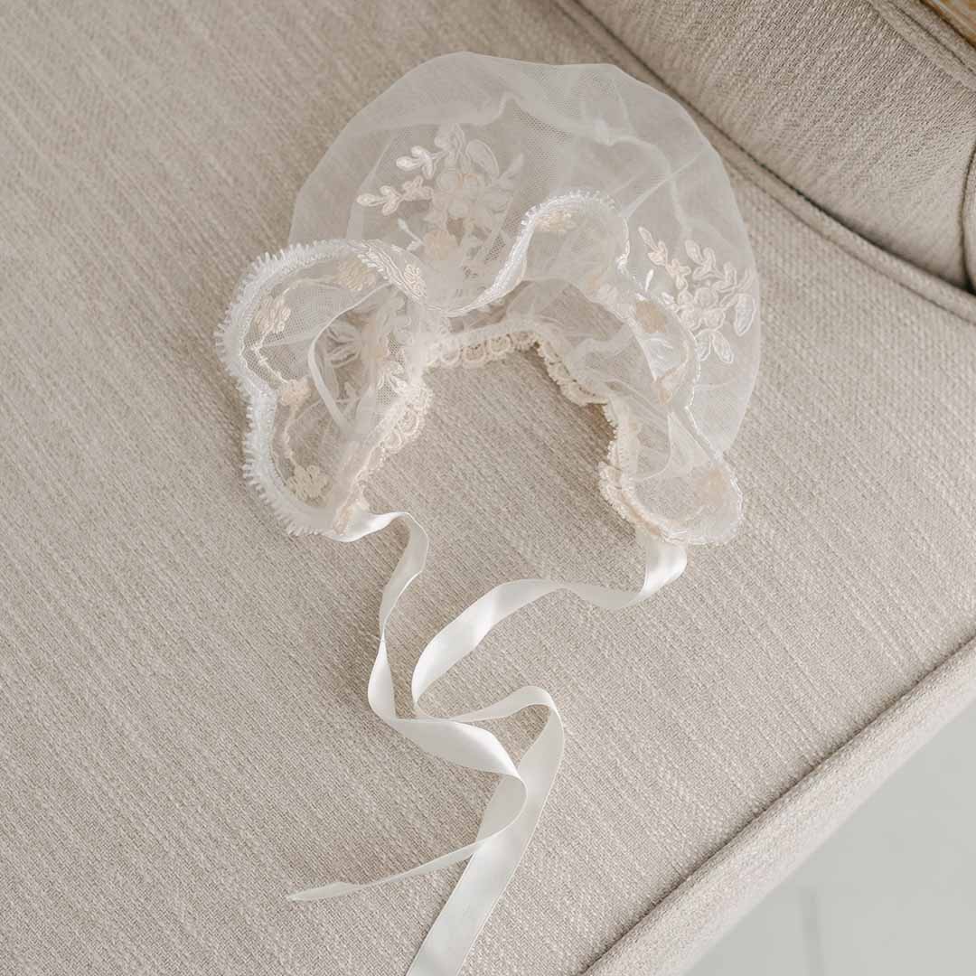 A delicate, vintage-inspired Kristina Christening Gown & Bonnet with floral embroidery and ribbon ties, resting on a soft beige upholstered chair.
