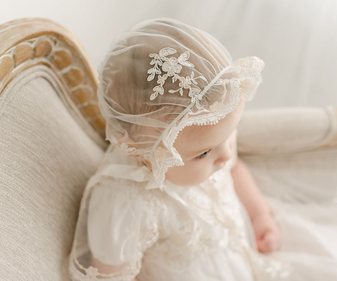 A close-up of a baby in the Kristina Christening Gown & Bonnet, sitting on a vintage sofa with a soft backdrop, ready for a christening.