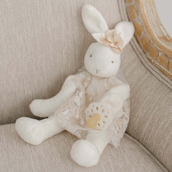 A Kristina Silly Bunny Buddy pacifier holder, an heirloom for a christening or baptism, with a lace dress and floral headband, sitting on a beige upholstered chair.