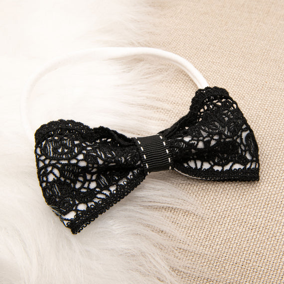 A black floral lace June Lace Bow Headband with a delicate floral pattern and a central metal clasp, displayed on a soft, beige textured background.