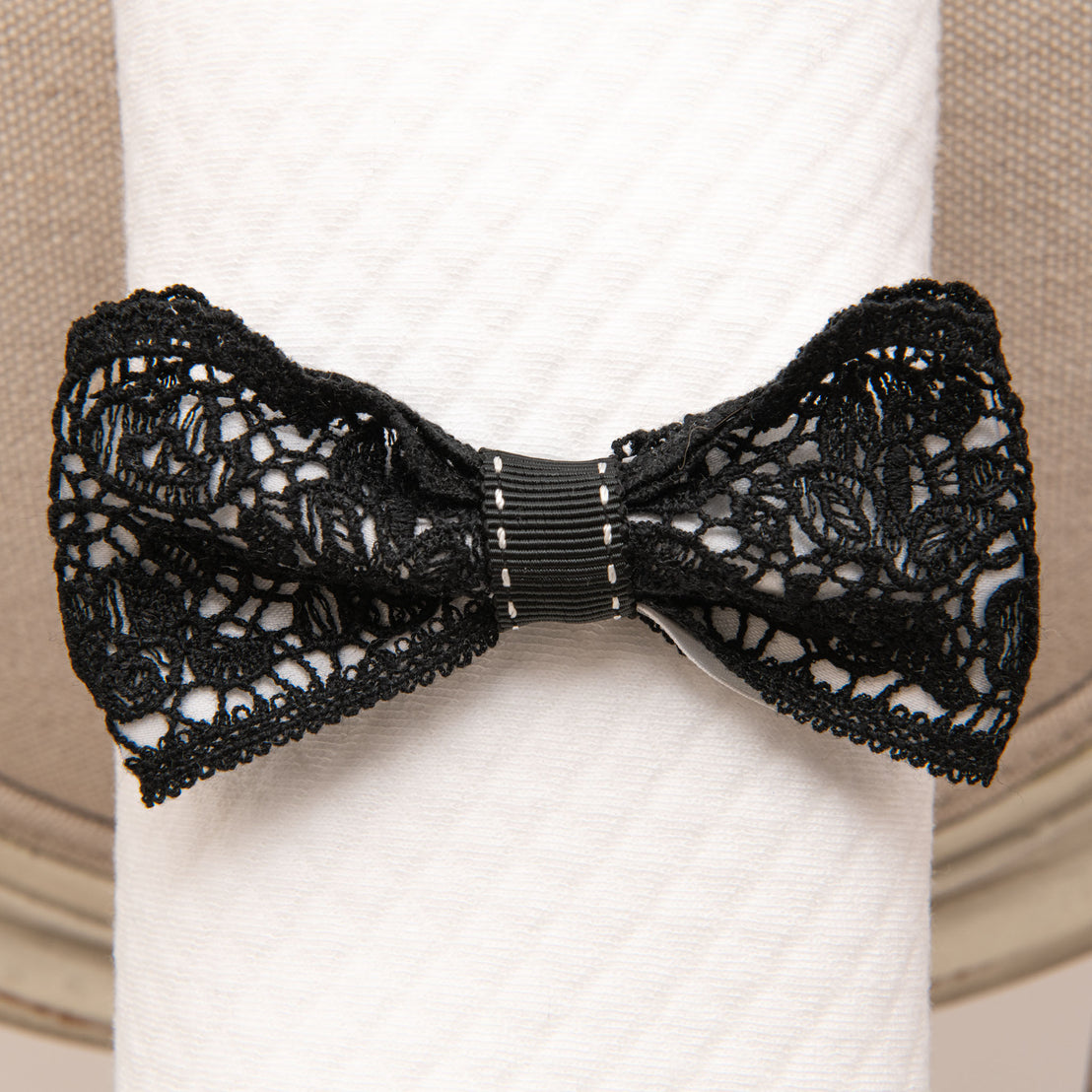 A decorative black floral lace bow tied around a white baby blanket, featuring intricate patterns and a small black clasp at the center. 