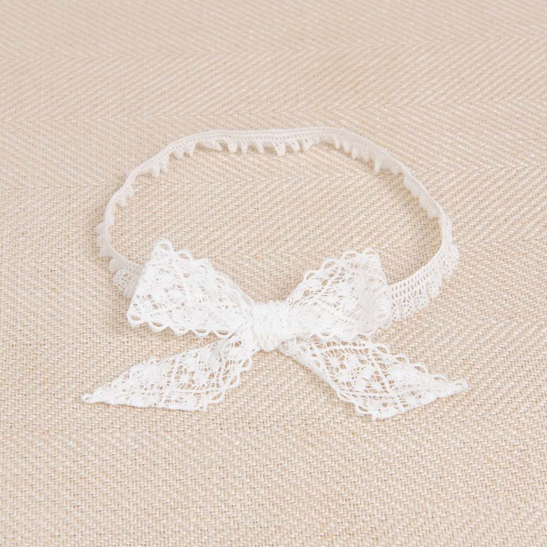A Thea Lace Headband, a delicate lace bow with light ivory stretch lace, displayed on a textured beige fabric background.