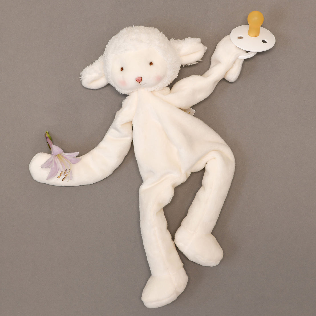 A Silly Lamb Buddy plush toy with a white fleece and limbs designed for an upscale baptism, lying on a grey background alongside a pacifier and a small purple flower.
