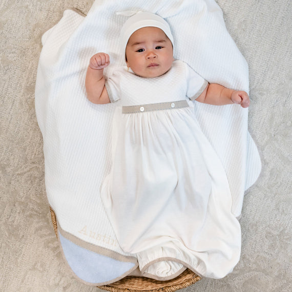 A baby dressed in the Austin Layette & Hat outfit and bonnet lies on a soft blanket, placed inside an upscale woven basket, looking up with a slight frown.