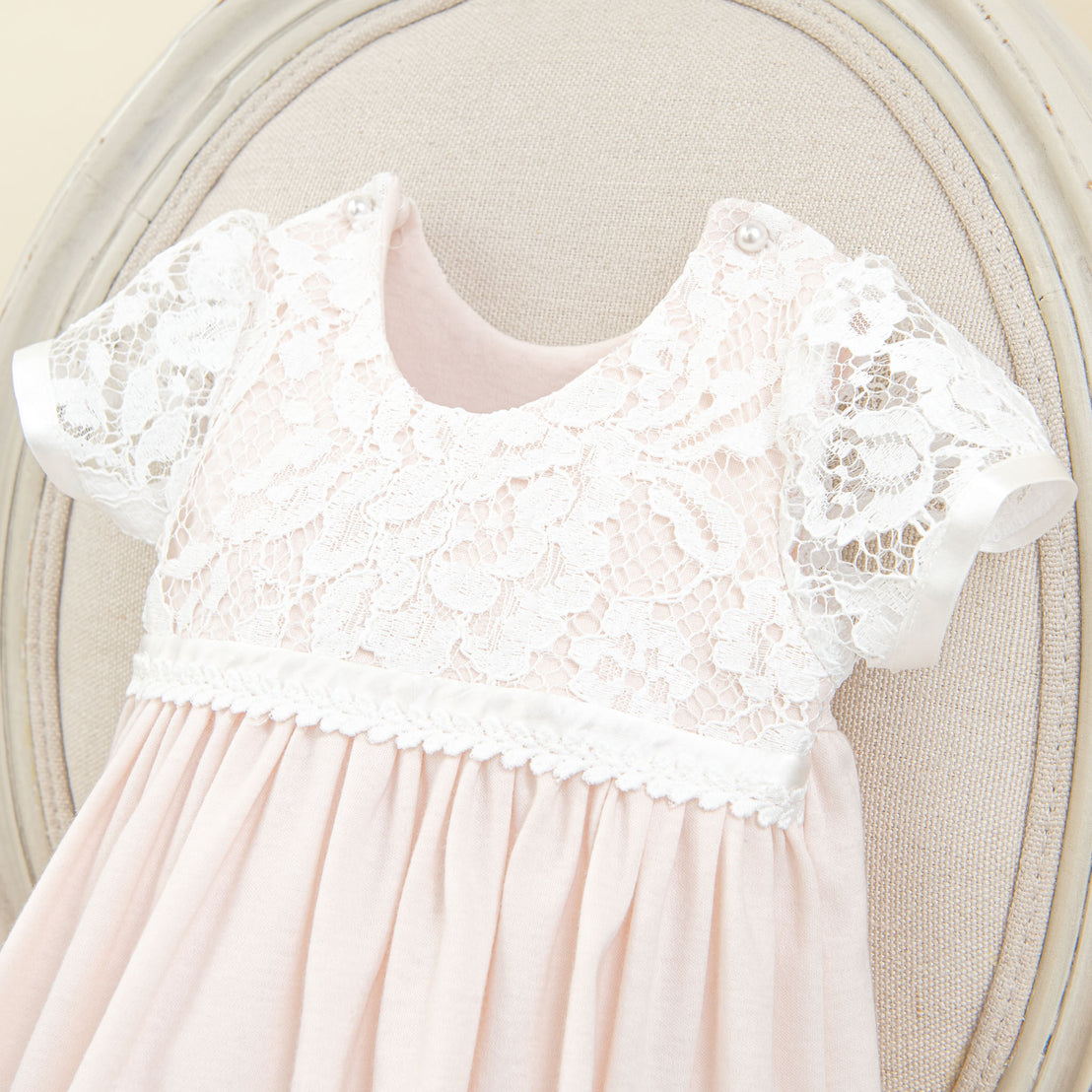 A pink Rose Layette & Bonnet dress with a delicate white lace upper and traditional cap sleeves, displayed elegantly as an heirloom on a beige antique chair.
