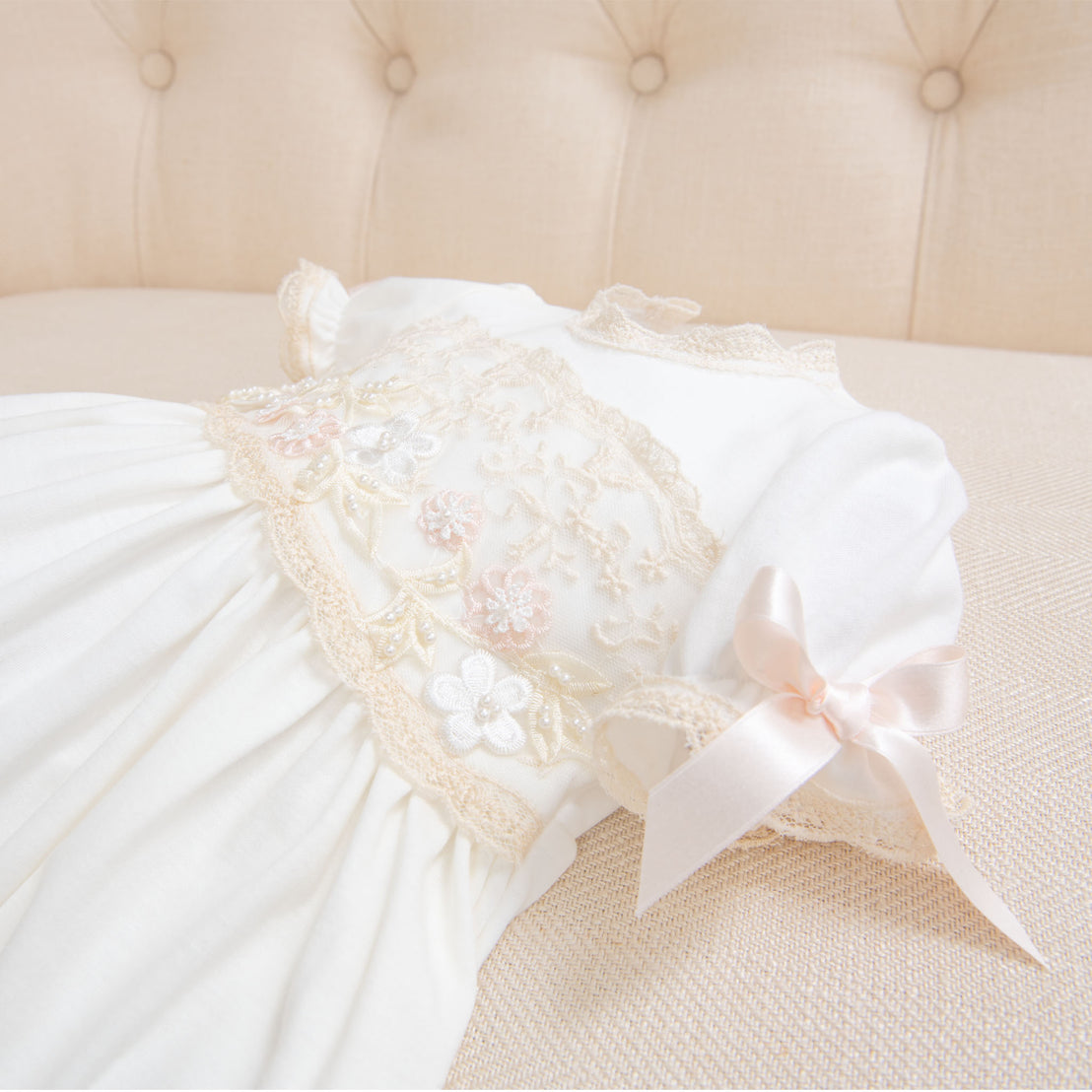 A close-up of the bodice on the Jessica Newborn Gown featuring delicate lace and floral embroidery, adorned with a pink bow on the sleeves, displayed on a soft beige couch.