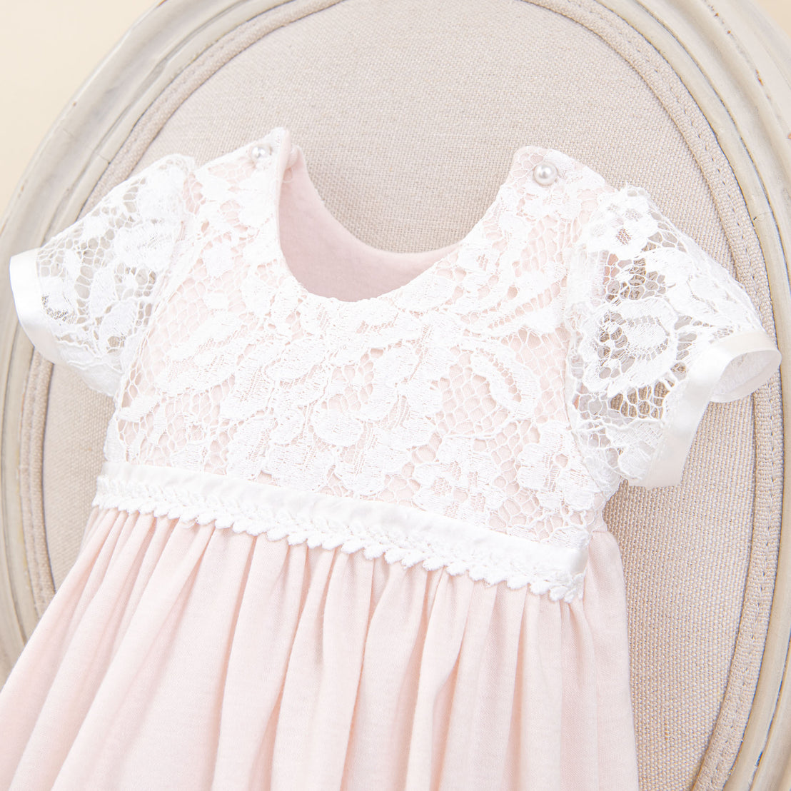 A delicate traditional Rose Newborn Layette with a white lace bodice and soft pink pleated skirt, displayed against a neutral beige circular backdrop.