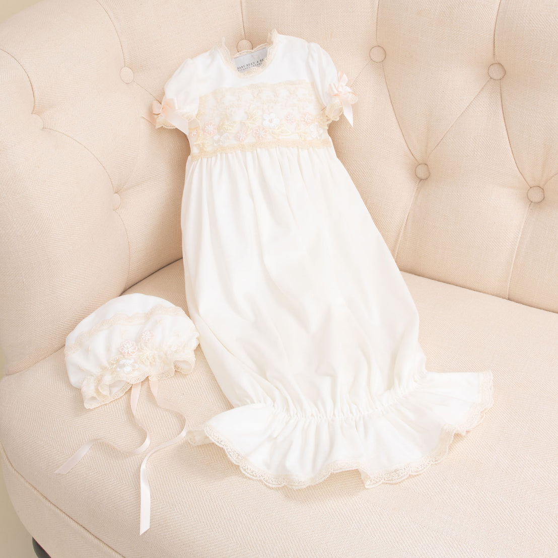 An elegant Jessica Newborn Gown with floral embroidery and lace details, accompanied by the matching Jessica Newborn Bonnet, laid out on a soft beige armchair.