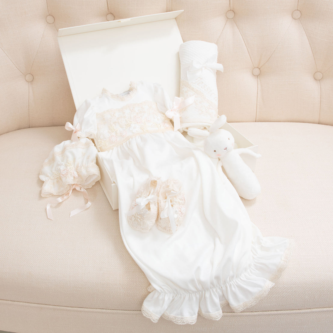 The items included in the Jessica Newborn Gift Set laid out of the white gift box on a beige sofa. The Jessica Newborn Gown is laid out along with the matching bonnet, booties, white bunny chime, and personalized blanket.