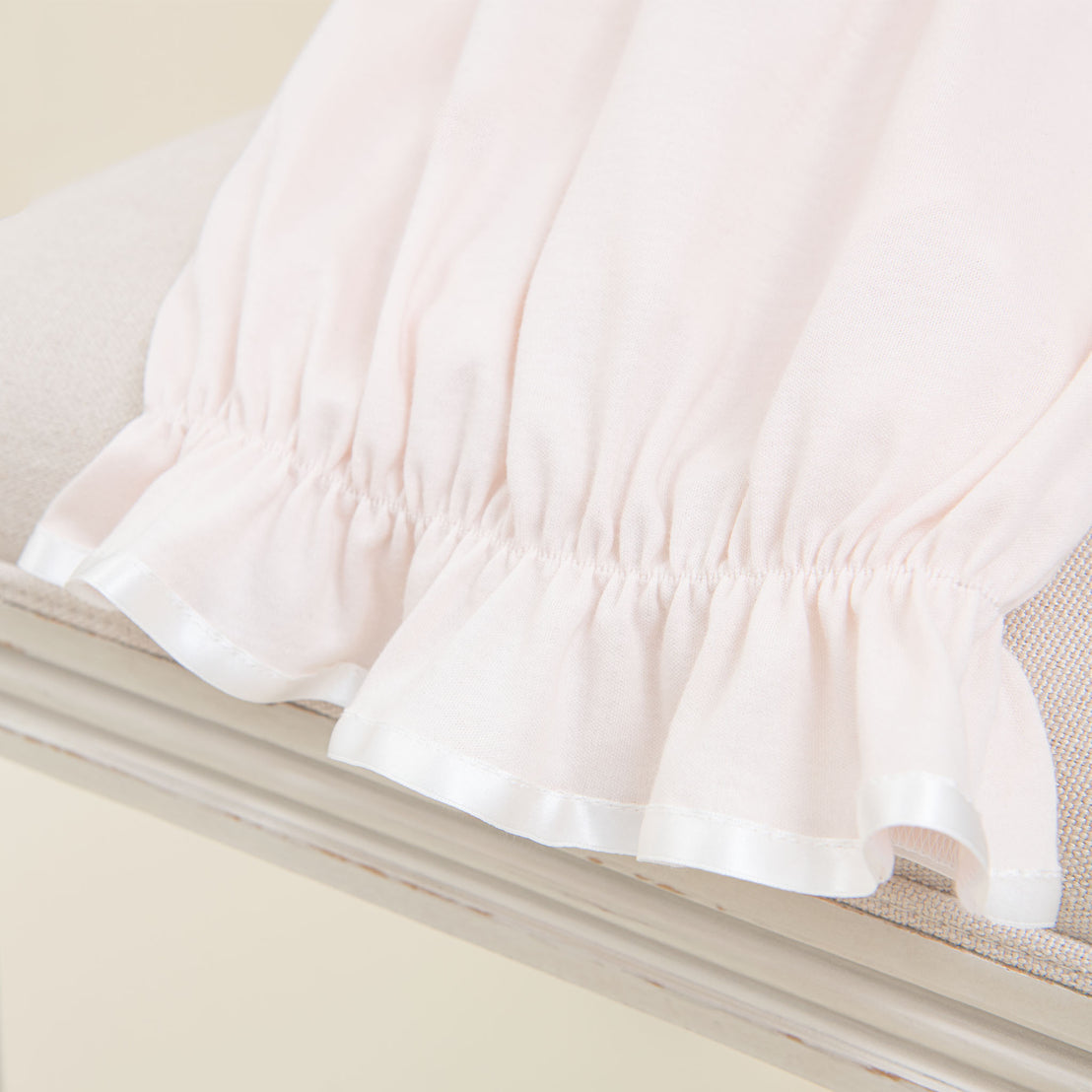 Close-up of a Rose Layette & Bonnet with a traditional light pink, ruffled edge lying on a beige surface, highlighting the delicate fabric texture and soft color tone.