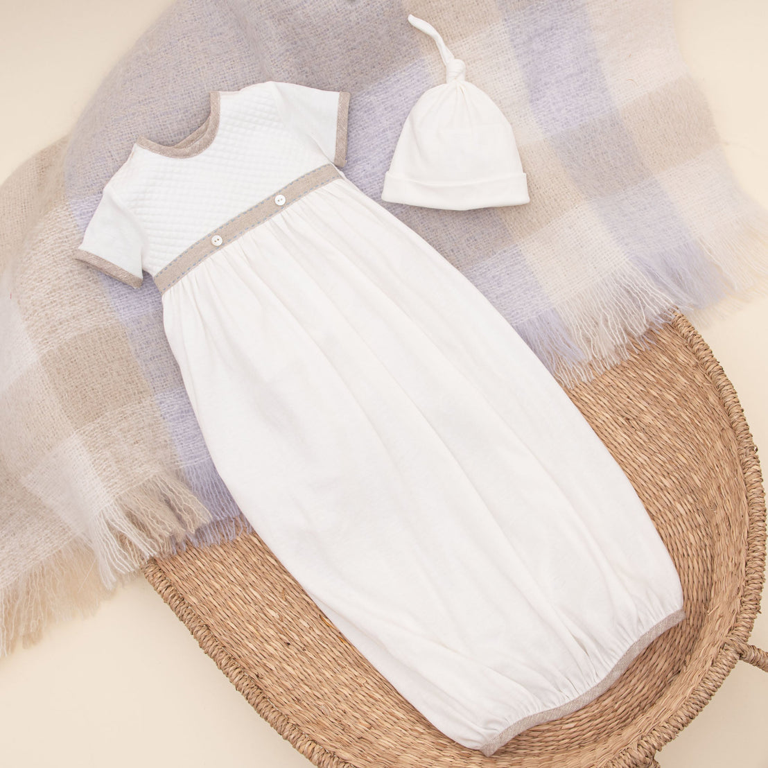 An Austin Layette & Hat, an heirloom-quality white gown with a textured upper bodice and grey details, is elegantly laid out on a wicker basket with a beige blanket and a matching.
