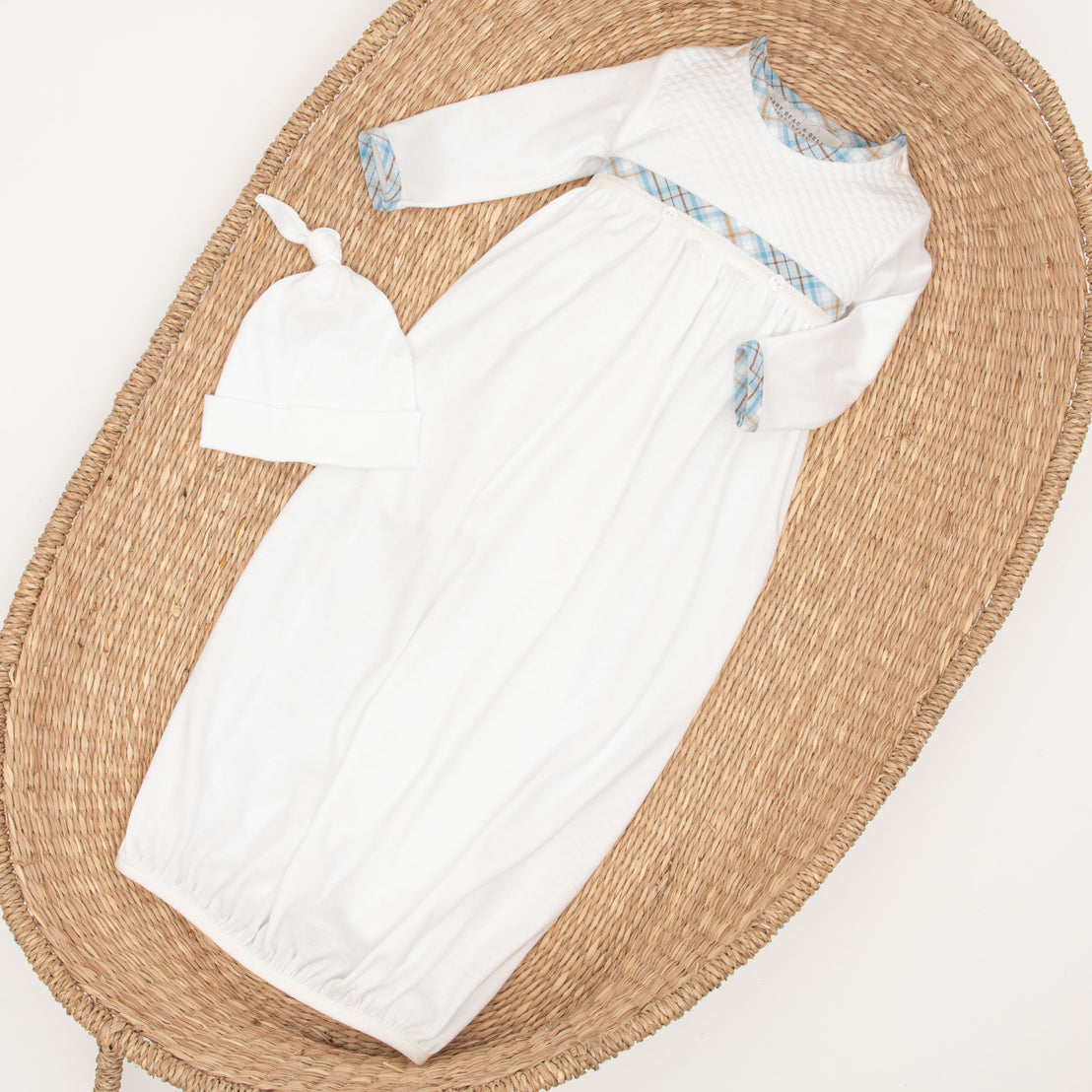 An upscale Mason Newborn Gift Set, consisting of a vintage white baby gown and matching hat with blue detailing, displayed on a round woven mat against a light background.