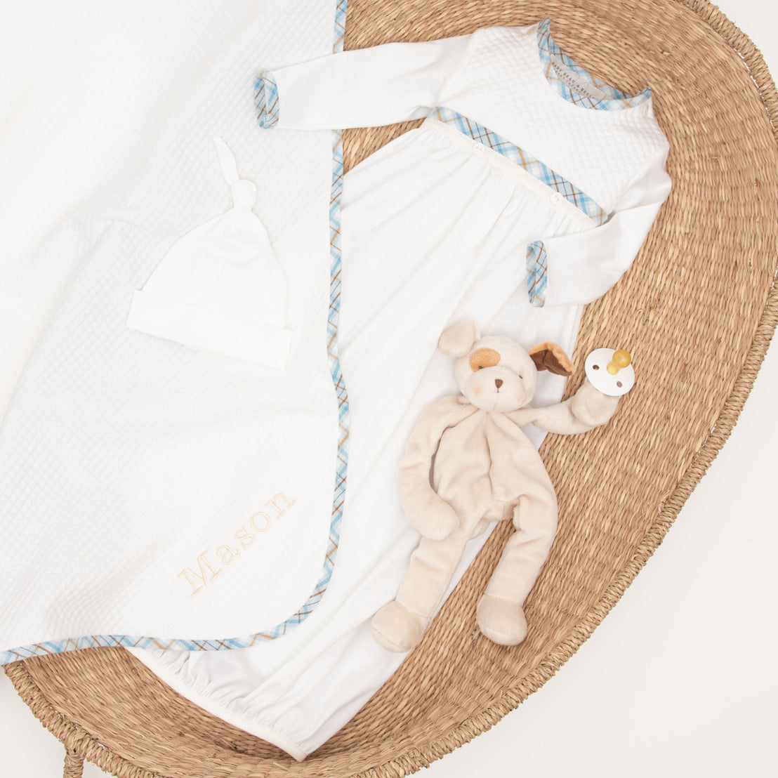 A baby's Mason newborn gift set comprising a white onesie, hat, and bib, personalized with the name "Mason", displayed with a plush puppy toy on an upscale woven round mat.