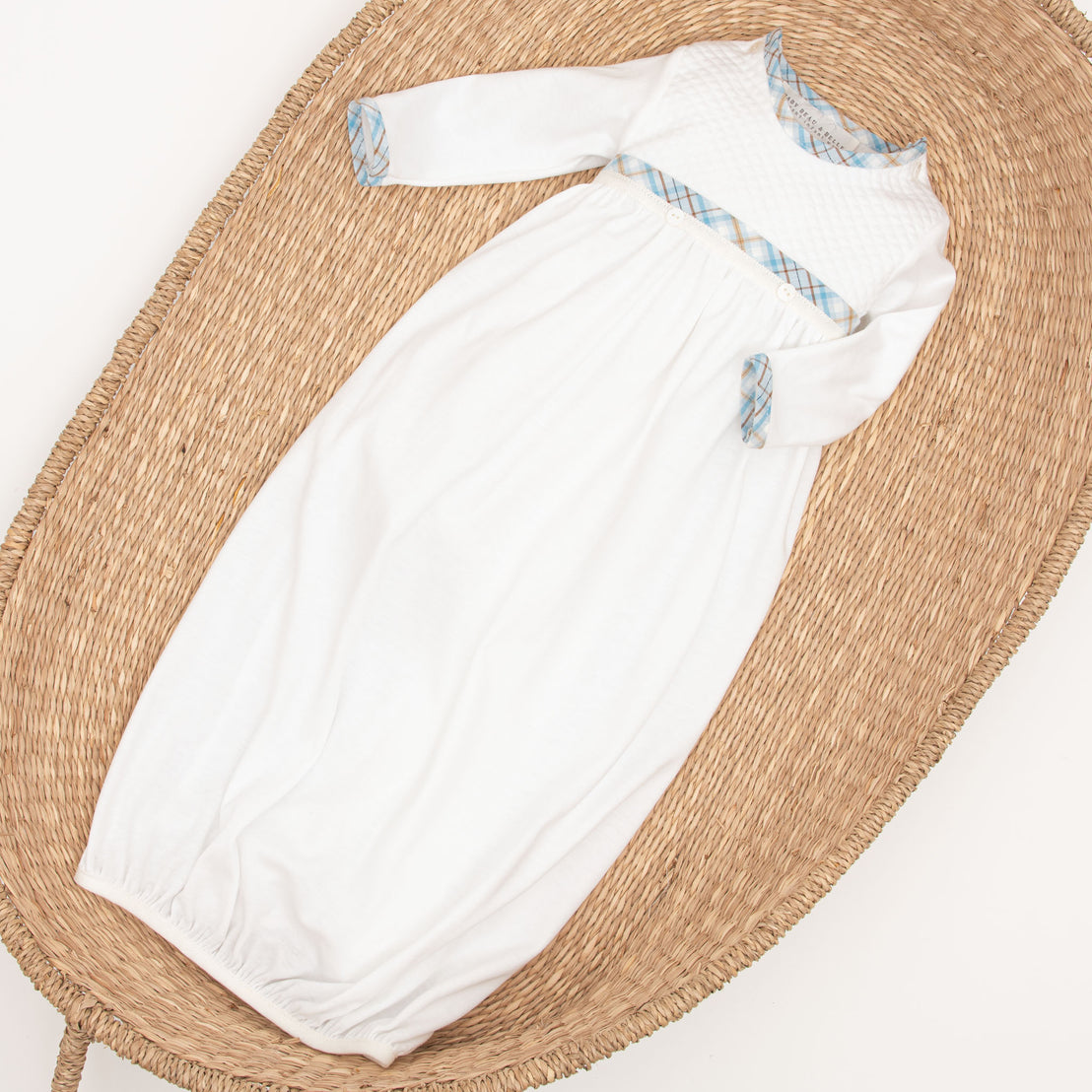 A white Mason Newborn Gown with blue trim laid neatly inside a woven, oval-shaped basket on a plain background.