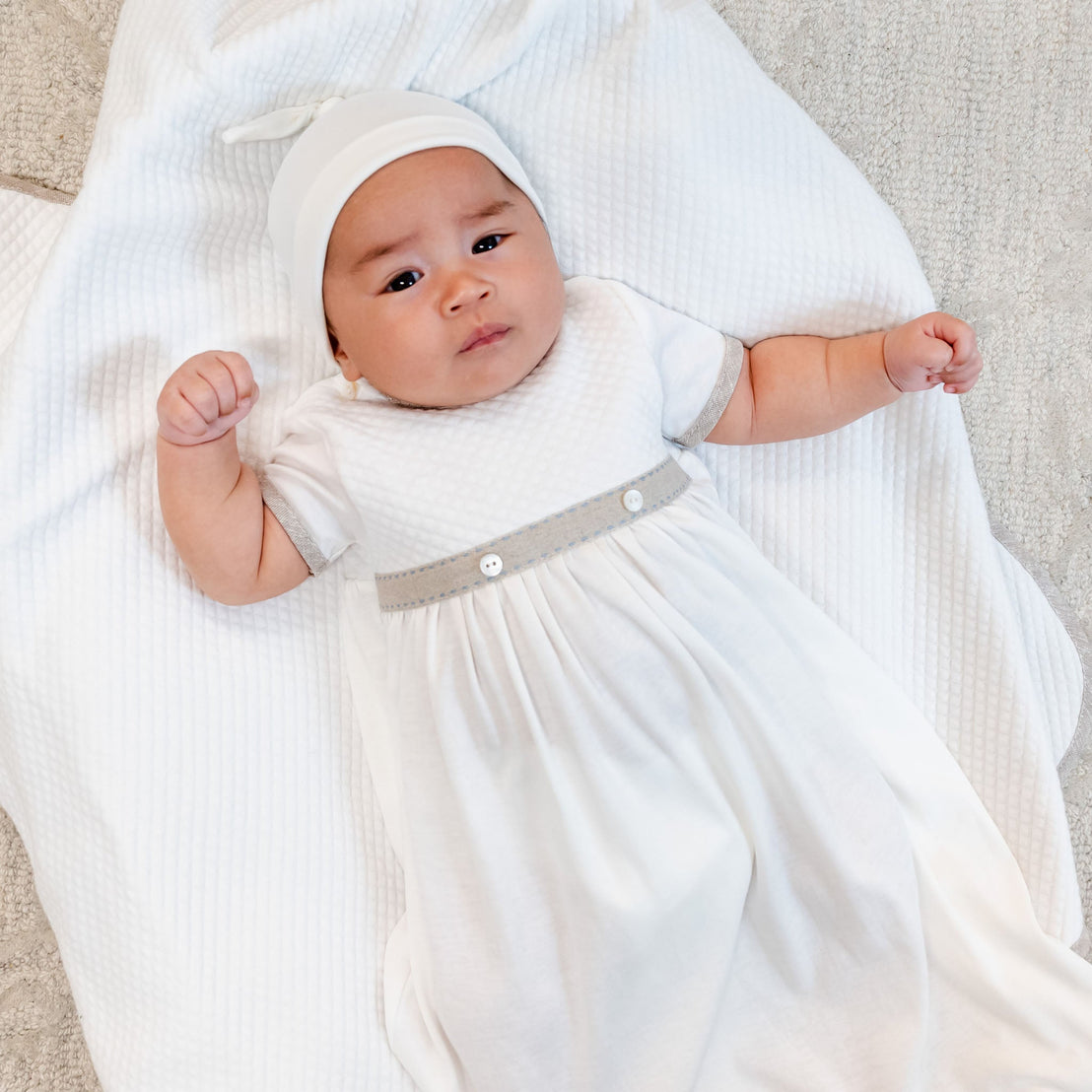 A baby dressed in the Austin Layette & Hat, white and gray coming home outfit lies on a soft blanket, looking upwards with a cute expression and arms outstretched.