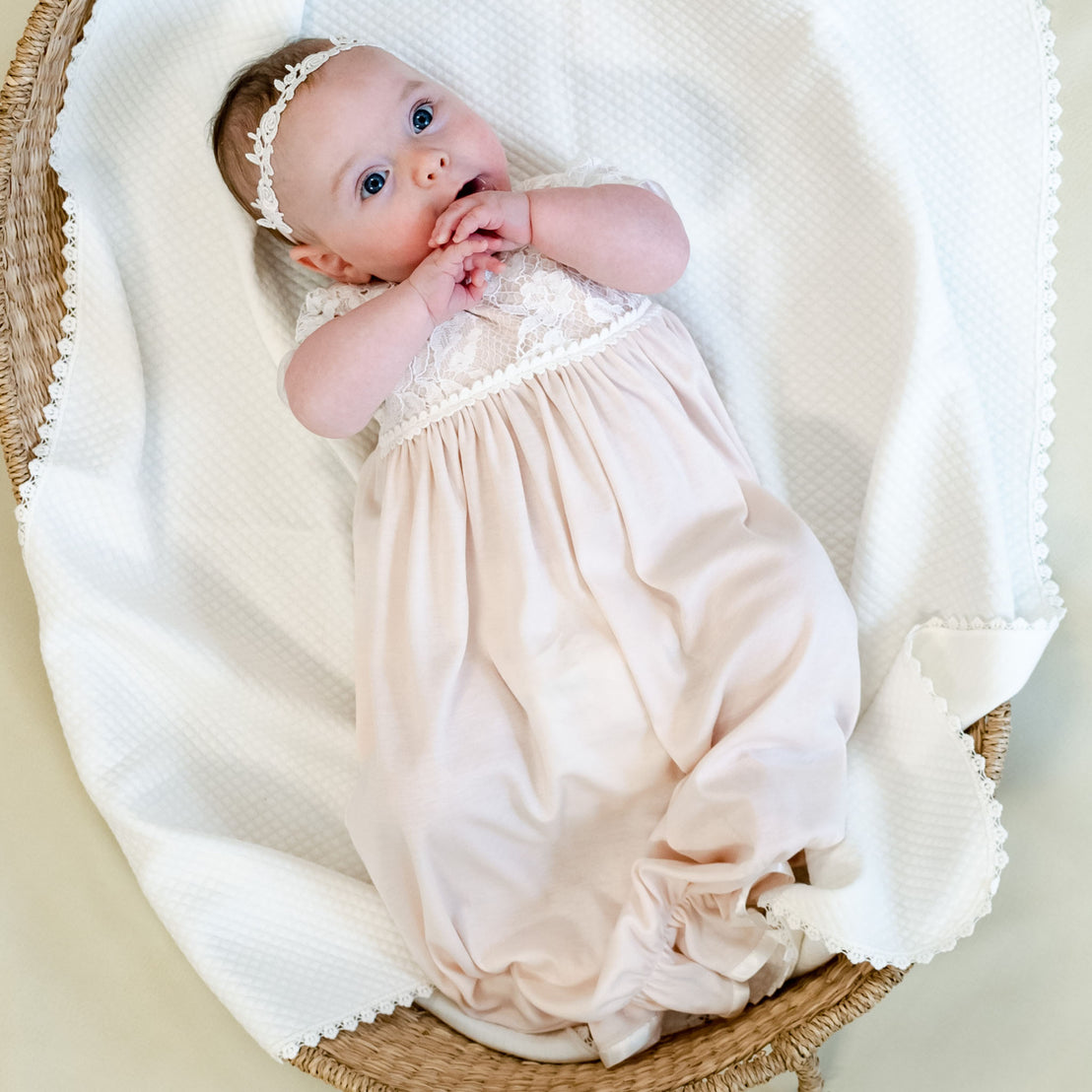A baby wearing a Rose Layette & Bonnet lies in a woven basket, looking upwards with hands clasped near the mouth, on a soft beige background.
