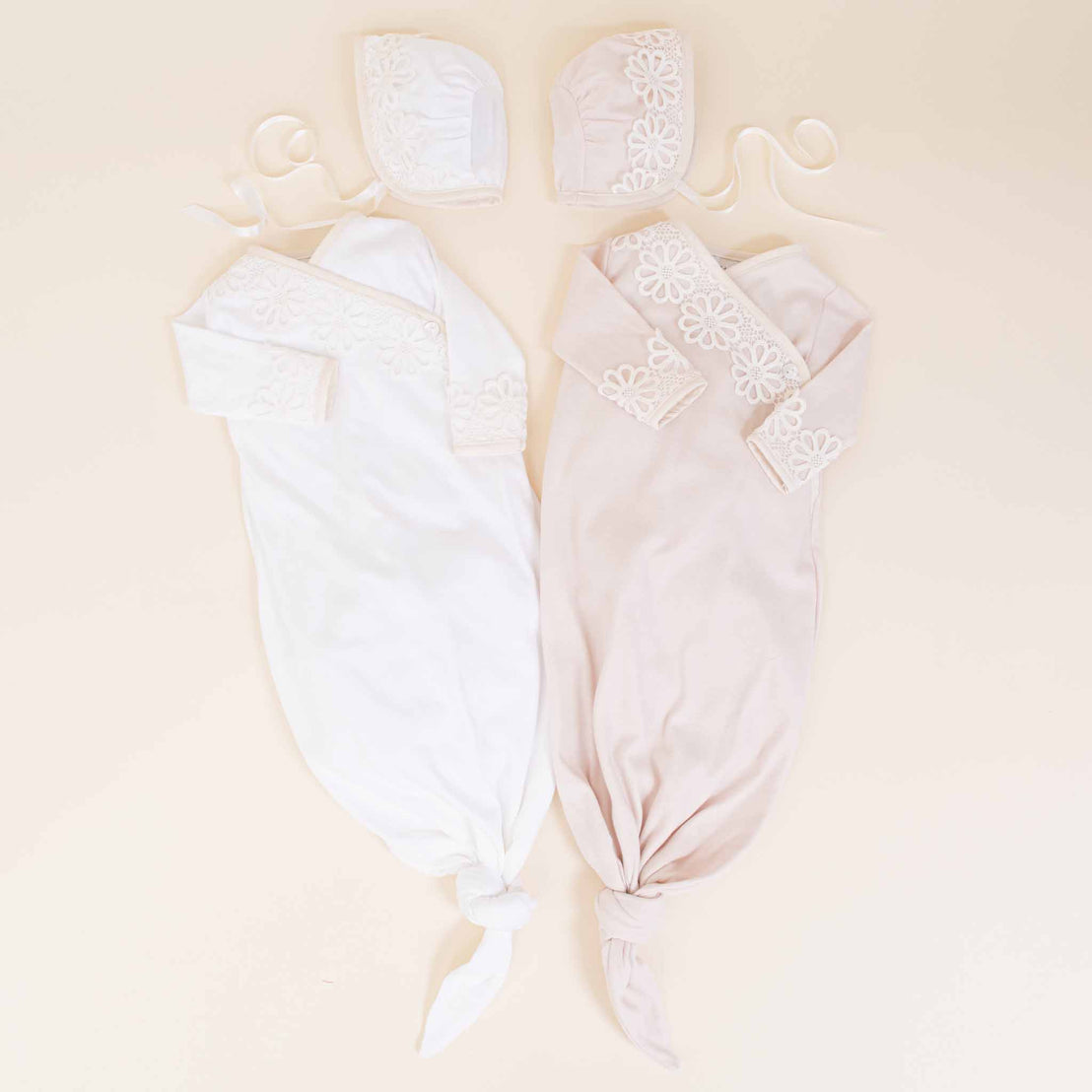 Two Hannah Knot Gowns & Bonnets by Baby Beau & Belle, displayed on hangers against a light beige background. One outfit is white and one is pastel pink, both adorned with lace detailing.