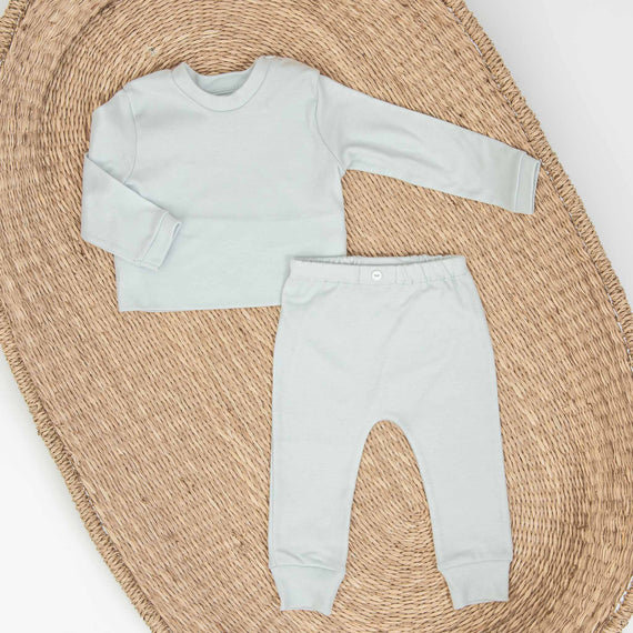 A mint green Aiden Pima Top & Leggings baby outfit comprising a long-sleeved top and pants neatly displayed on a woven circular mat. The top has a round neckline and the upscale pants include a button at the waistband.