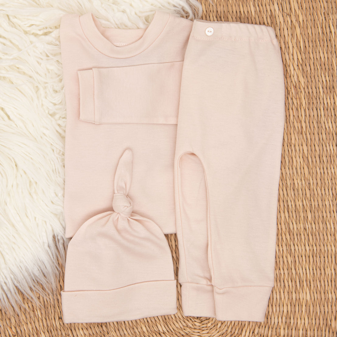 The blush pink Ava Pima Top & Leggings coming home outfit including a long-sleeved pima cotton shirt, pants, and matching knotted cap, displayed folded on a wicker basket with a white fluffy rug beneath.