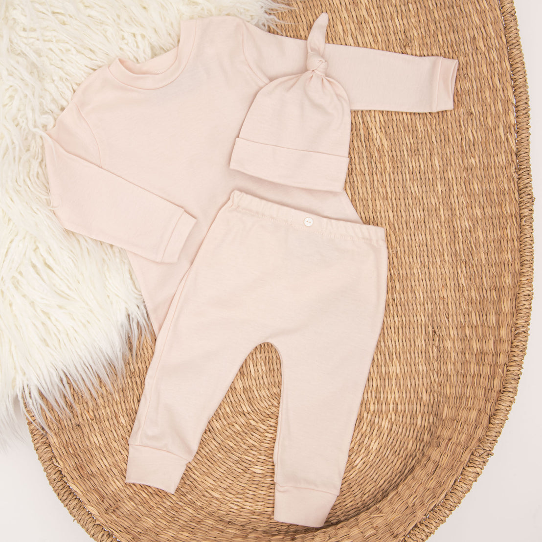 A flat lay of a baby's light pink coming home outfit including theAva Pima Top & Leggings set, and matching Ava Pima Knot Cap, arranged on a wicker basket with a white fluffy rug beneath.