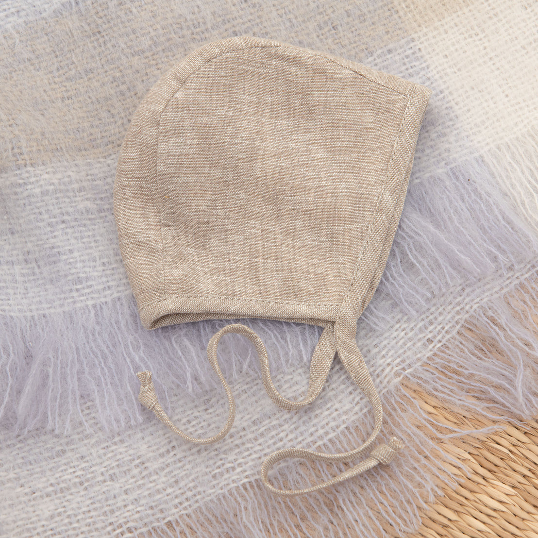 An upscale Austin Linen Bonnet with ties, placed on a soft, lilac textile background, perfect for a coming home outfit.