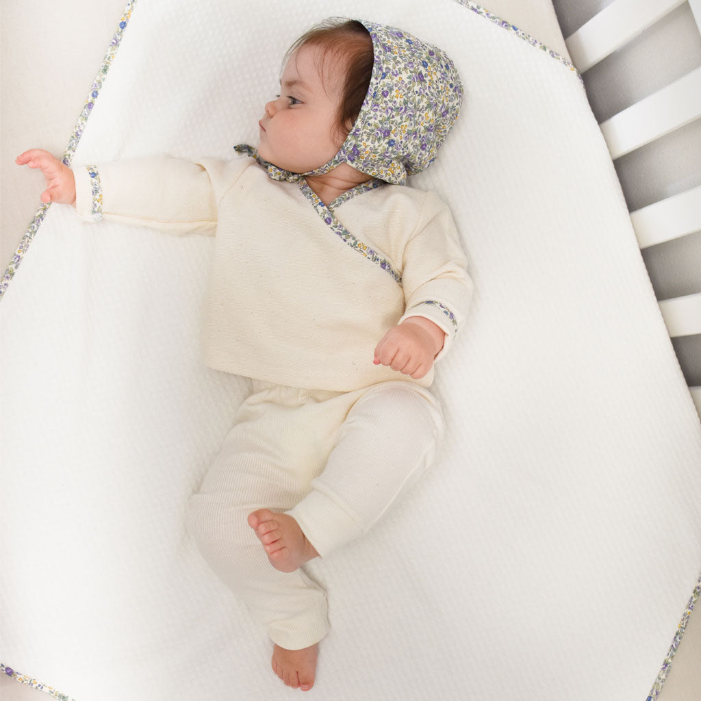 A baby wearing a boutique floral bonnet and heirloom cream outfit lies peacefully in a white crib with grey bars, one arm stretched out next to the Petite Fleur Personalized Blanket.