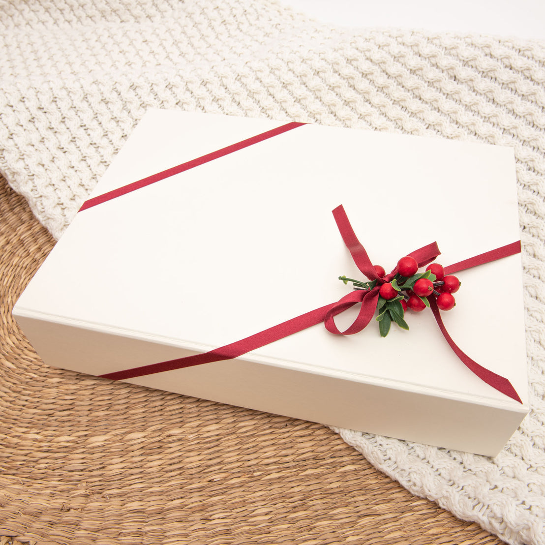 A rectangular ivory gift box with a magnetic closure, tied with a red ribbon and decorated with a small cluster of red berries and green leaves, resting on a knitted cream blanket with a woven background.