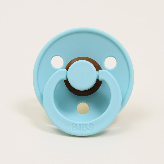 A mint green pacifier with a round brown handle and the brand name "bibs" written at the top, designed in Denmark, set against a plain white background.