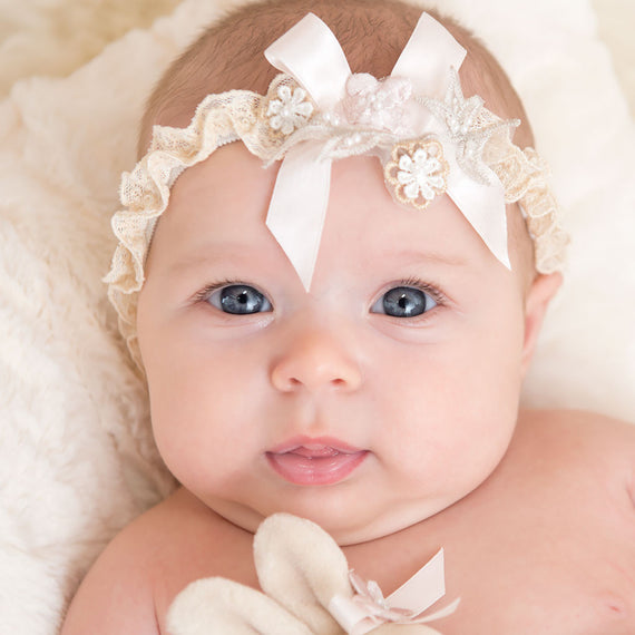A close-up of a baby with bright blue eyes wearing a Jessica Headband with a bow, nestled in a soft, white blanket. The baby appears calm and is looking directly at the viewer.