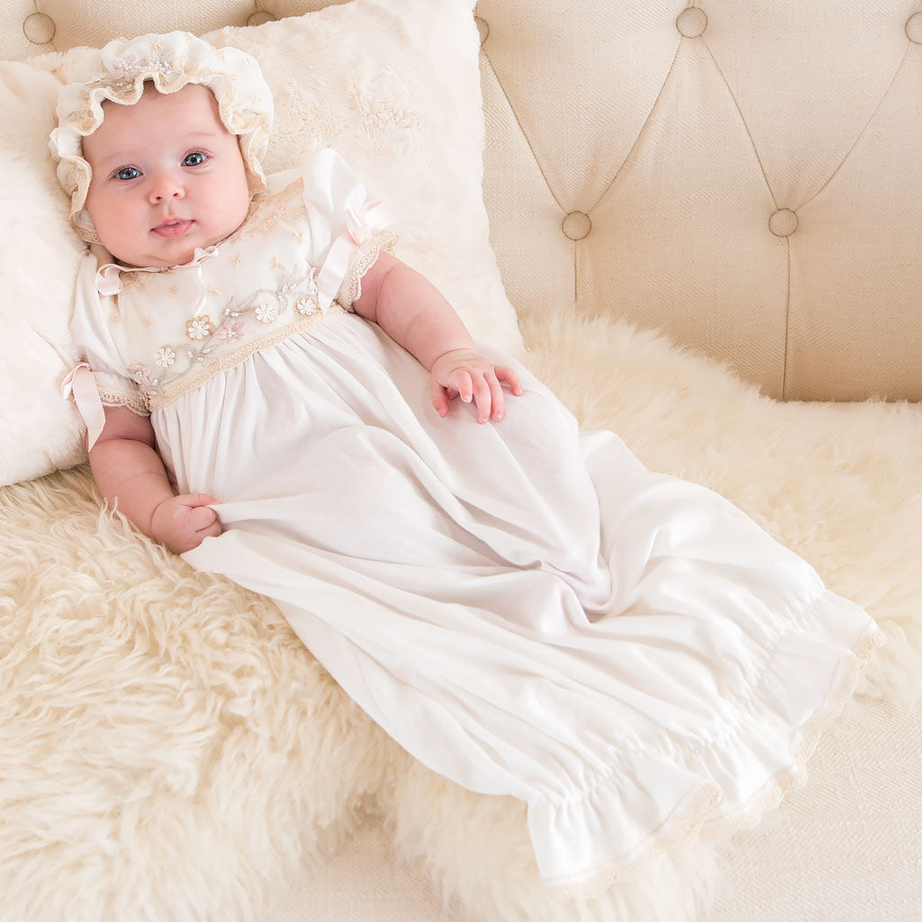 A baby in a Jessica Newborn Gown & Bonnet lies on a fluffy white blanket on a beige couch, looking at the camera with a slight smile.